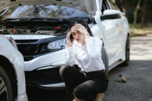 Woman crouching beside damaged car with open hood while speaking on phone